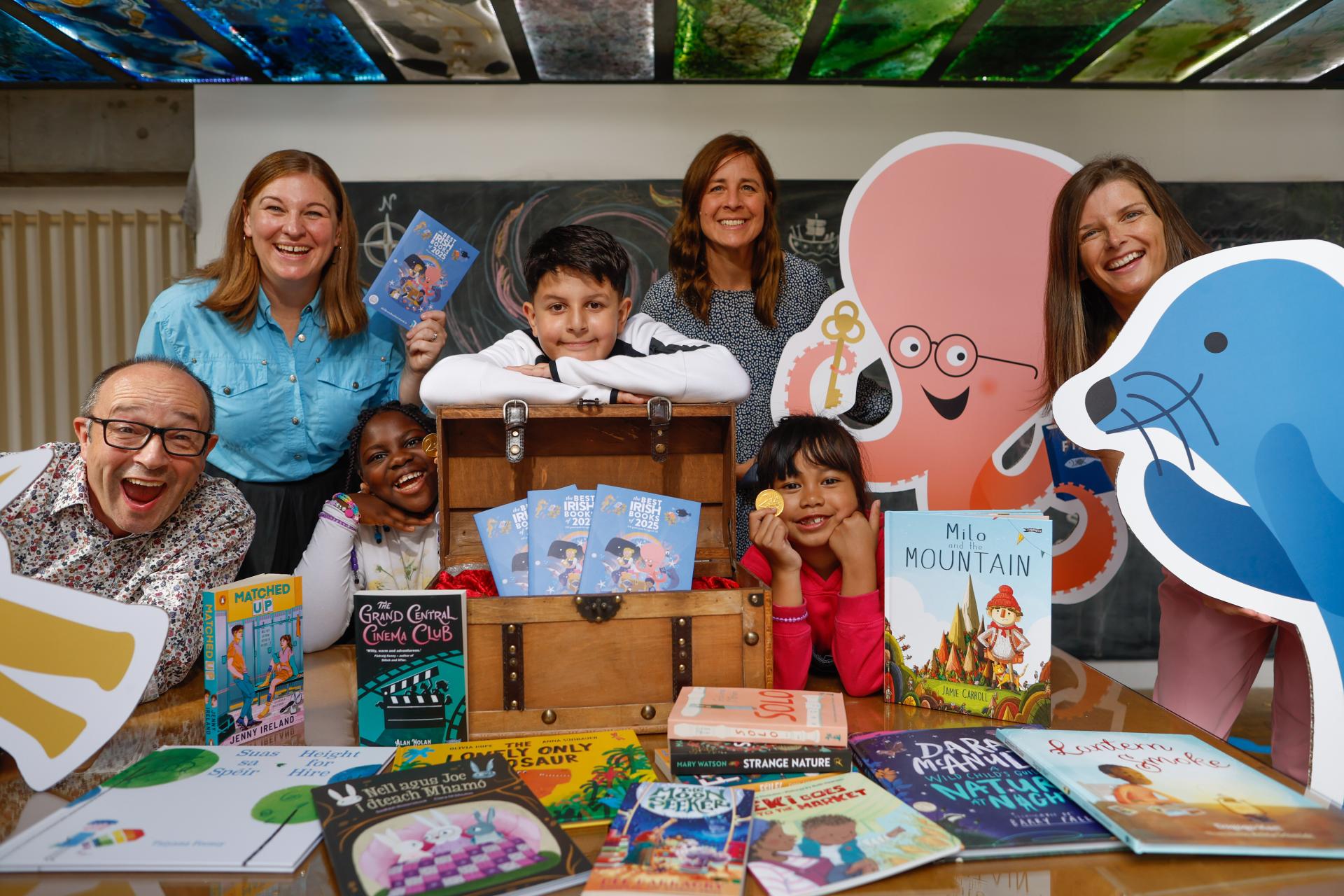 Authors, illustrators, students and CEO of CBI celebrate in the launch of The Best Irish Books, gathered around a table with a treasure chest, gold coins and a selection of books