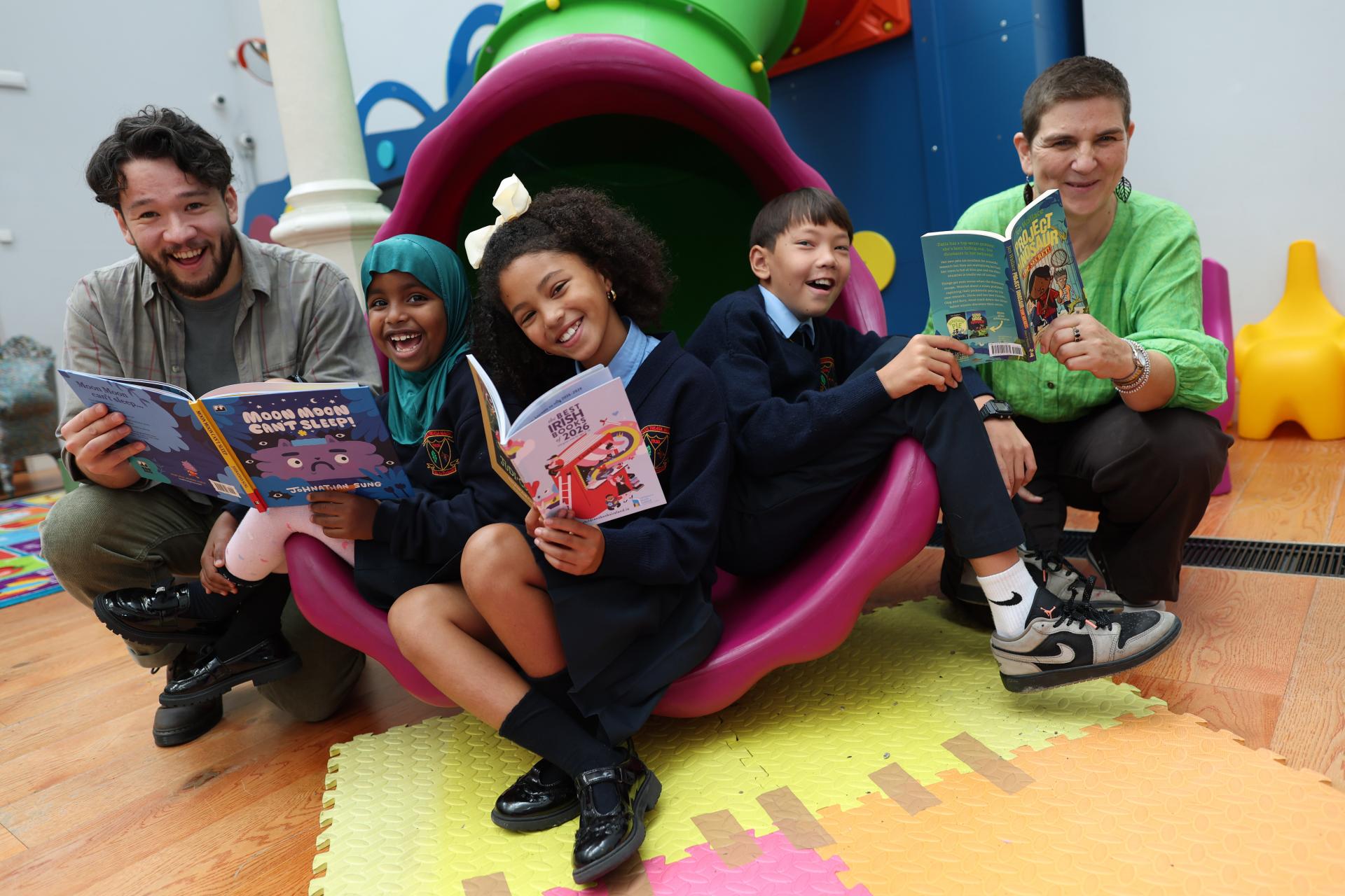 Two authors and three children sitting on the end of a slide in a library space. They are all holding up copies of books and the Children's Books Ireland reading guides.