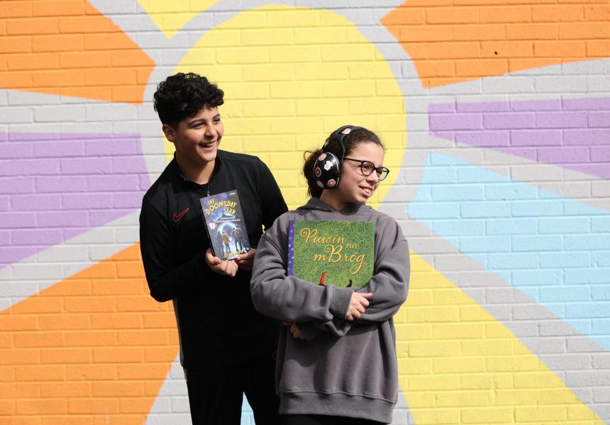 Two students stand in front of a mural of a sun holding books and staring off into the distance.